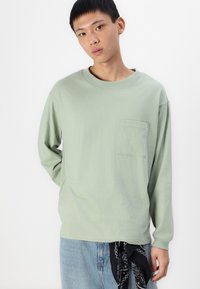 Mintgroene katoenen blouse met lange mouwen en een ronde halslijn, voorzien van een borstzak, gecombineerd met lichte spijkerbroeken en een zwarte bandana als accent.