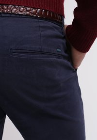 Mann trägt marineblaue Hosen mit Gesäßtasche, braunen strukturierten Gürtel und burgunderfarbenen Pullover, die Hand in der Tasche.