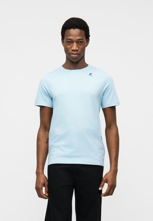 Άνδρας στέκεται μπροστά σε λευκό φόντο φορώντας ανοιχτό μπλε κοντομάνικη μπλούζα με μικρό λογότυπο στο στήθος και μαύρο παντελόνι.