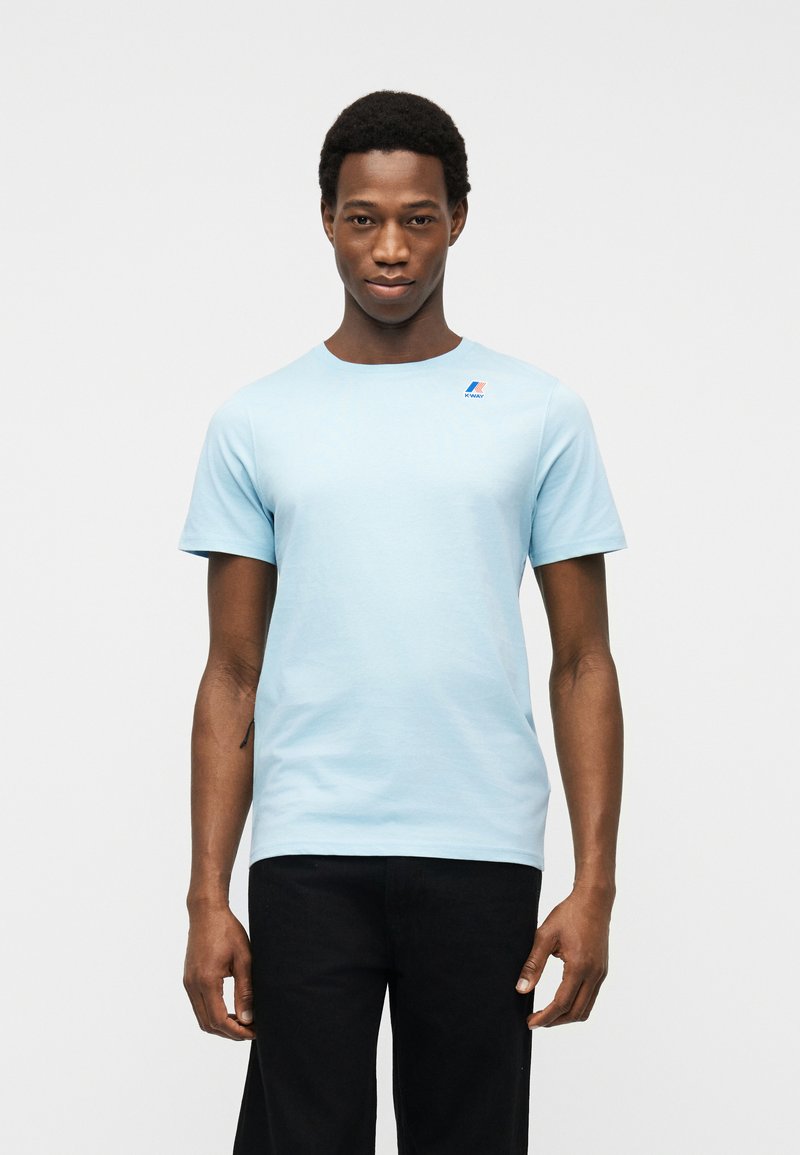 Άνδρας στέκεται μπροστά σε λευκό φόντο φορώντας ανοιχτό μπλε κοντομάνικη μπλούζα με μικρό λογότυπο στο στήθος και μαύρο παντελόνι.