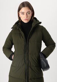 Olijfgroene pufferjas met een capuchon, gestikt ontwerp, rits aan de voorkant en zijzakken. Draagt over een zwarte turtleneck en vastgehouden met een zwarte tas.
