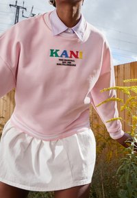 Sudadera rosa claro con puños y dobladillo acanalados, que presenta un logo "KANI" multicolor y texto en negro. Llevada sobre una falda blanca con pliegues.