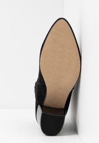 Chaussure en suède noir avec un embout pointu et une semelle en caoutchouc beige lisse. Le talon est modérément épais et incliné pour plus de stabilité.