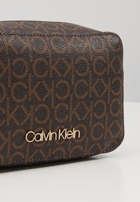 Coin de sac en cuir texturé marron avec un motif répété "ck" et le logo doré Calvin Klein sur une surface blanche.