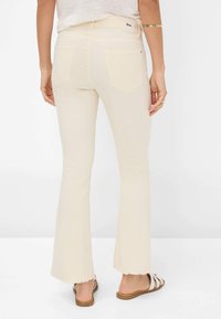 Crème kleurige flared broek met een rafelrand, twee achterzakken en een merklabel op de tailleband. Het materiaal lijkt glad en rekbaar.