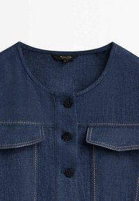 Veste en denim bleu foncé avec encolure ronde, trois boutons noirs et deux poches poitrine à rabat avec surpiqûres beige.