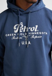 Felpa blu navy con logo bianco "Petrol", dettagli testuali e cordini con punte metalliche. Tessuto morbido, vestibilità casual.
