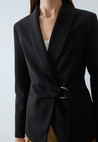 Mustat bleiseri, jossa on terävä läppä ja kapea vyötärö hopeisella soljella. Valmistettu sileästä kankaasta, jossa on säädetty istuvuus ja tyylikäs muotoilu.