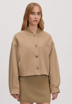 Beige Cropped-Jacke mit hohem Kragen, großen Knöpfen und einer Brusttasche; aus einem weichen Stoff mit glatter Textur gefertigt.