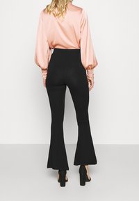 Svarta utsvängda byxor med en slät textur, hög midja och figurnära silhuett, tillsammans med en rosa satinblus med draperade ärmar.