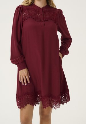 Femme portant une robe bordeaux à manches longues avec des détails en dentelle sur la poitrine et l'ourlet, ainsi que des poignets froncés.