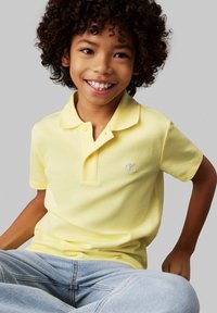 Bambino sorridente con capelli ricci, che indossa una maglietta polo gialla e jeans azzurri chiari, seduto contro uno sfondo semplice.