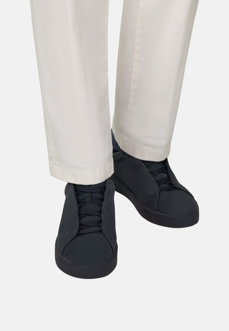 Personne portant des baskets noires à lacets et un pantalon beige clair, debout les pieds près l'un de l'autre sur un fond uni.
