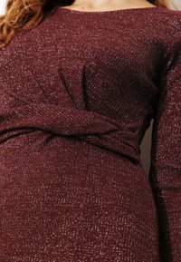 Robe bordeaux à manches longues, avec une finition texturée et un détail de taille nouée. Des fibres scintillantes ajoutent une subtile brillance.