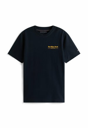 Camiseta negra de manga corta con pequeño texto amarillo "The Hilliger Herald MCMLXXXV" en el lado izquierdo del pecho y un pequeño logo en la manga derecha.