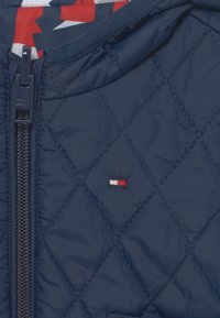 Námořnicky modrá prošívaná bunda s uzavíráním na zip, elastickým límcem a malým vyšívaným logem v červené, bílé a modré barvě na přední straně.
