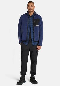 Marinblå fleecejacka med svarta detaljer, dragkedja fram, sidofickor, kombinerad med en blå rutig skjorta och svarta cargobyxor. Rejäla svarta skor.