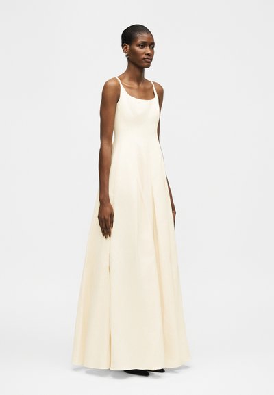 Γυναίκα που φοράει αμάνικο, μακρύ μέχρι το πάτωμα, κρεμ φόρεμα με εφαρμοστό μπούστο και φουσκωτή φούστα, στέκεται μπροστά σε ένα απλό ανοιχτό φόντο.