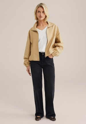 Giacca leggera beige con colletto rialzato, chiusura a zip e polsini elastici, abbinata a jeans a gamba larga di colore scuro e scarpe con stampa leopardata.