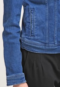 Modrá džínová bunda s přiléhavým střihem, vybavená zipovým zapínáním na přední části, bočními kapsami a kontrastním prošíváním podél švů.