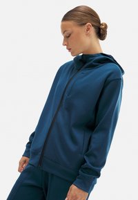 Sudadera azul de tejido suave con cierre de cremallera frontal, cuello alto y mangas largas. Presenta un ajuste holgado y sin patrones o elementos metálicos visibles.