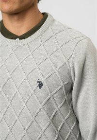 Šviesiai pilkas megztinis su deimantine mezgimo raštu, raišteliu aplink kaklą ir mažesniu siuvinėtu logotipu ant krūtinės. Pagamintas iš tekstūruotos medžiagos.