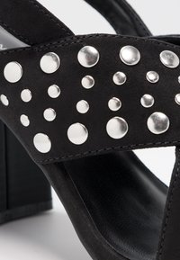 Sandales à talons hauts en daim noir avec une large lanière ornée de clous métalliques argentés de différentes tailles. Design de talon carré.