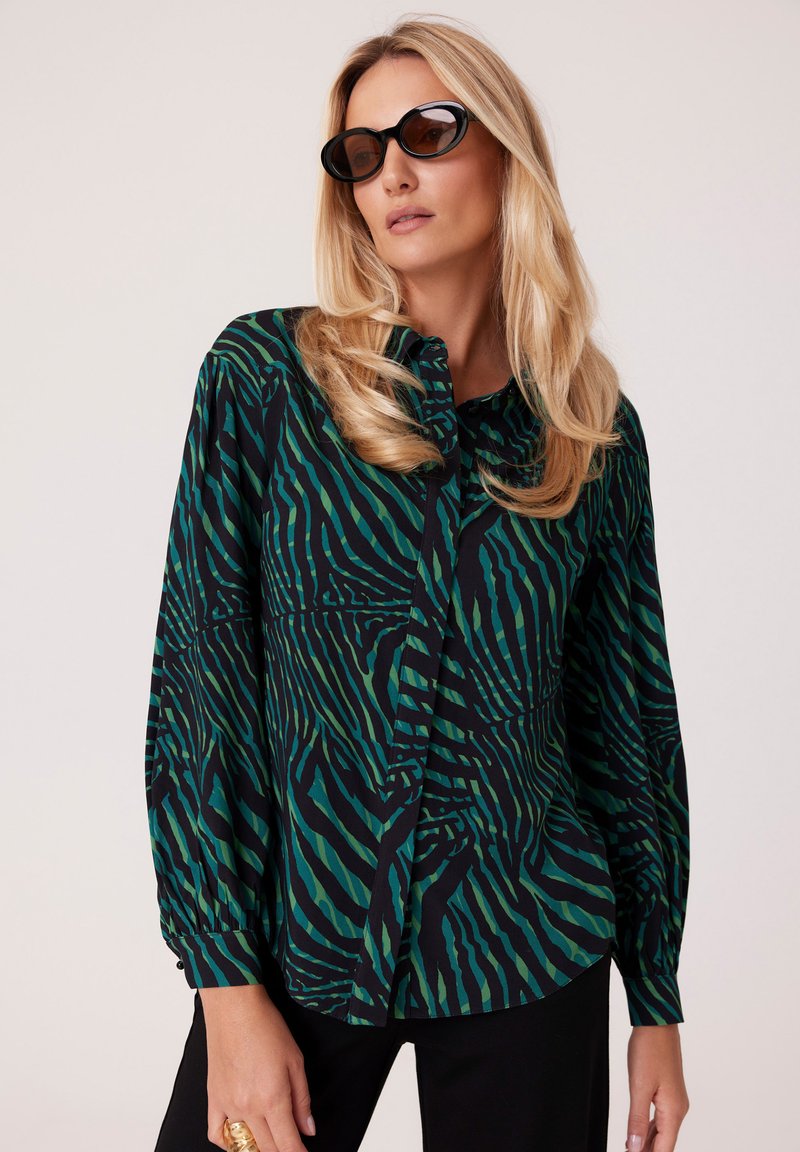 Zielono-czarna bluzka w wzory z długimi rękawami, z zapięciem na guziki z przodu, wykonana z miękkiego materiału; w połączeniu z czarnymi spodniami i dużymi okularami przeciwsłonecznymi.