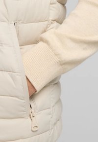 Chaleco acolchado de color beige claro con textura de acolchado, que cuenta con un bolsillo lateral con cremallera y un suave puño de suéter beige visible debajo.