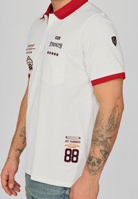 Vit polotröja med röd krage, framficka och olika broderade detaljer inklusive siffror och text. Korta ärmar och lätt material.