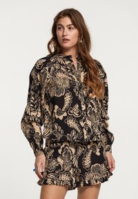 Blusa nera con motivo floreale beige, caratterizzata da un colletto alto e un'apertura con bottoni. Abbinata a pantaloni corti coordinati con orlo arricciato.