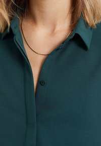 Teal knapp-up skjorta med krage, med en slät tygtextur och en smal knappslå. En tunn guldhalsband syns vid halsen.