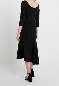 Svart knälång klänning med 3/4 långa ärmar och rak rygg. Mjuk, flödande tyg. Bärs med svarta ankelsandaler med rem. Enkelt och elegant design.