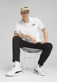 Polo bianca con logo nero, pantaloni neri, cappello beige e sneakers bianche con dettagli neri, seduto su una sedia di filo bianco.