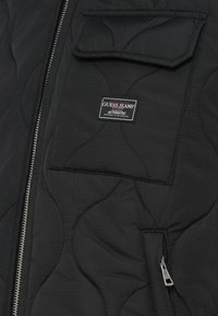 Juodas siuvinėtas paltas su užtrauktuku, krūtinės kišene ir logotipu. Yra tekstūruotas audinys su išlinkimo siūlėmis.