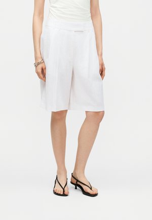 Γυναίκα που φοράει λευκή ψηλόμεση σορτς με φαρδιά πόδια μέχρι το γόνατο, λευκό αμάνικο τοπ, μαύρα σανδάλια με λεπτά λουράκια και ασημένιο βραχιόλι στον αριστερό καρπό.