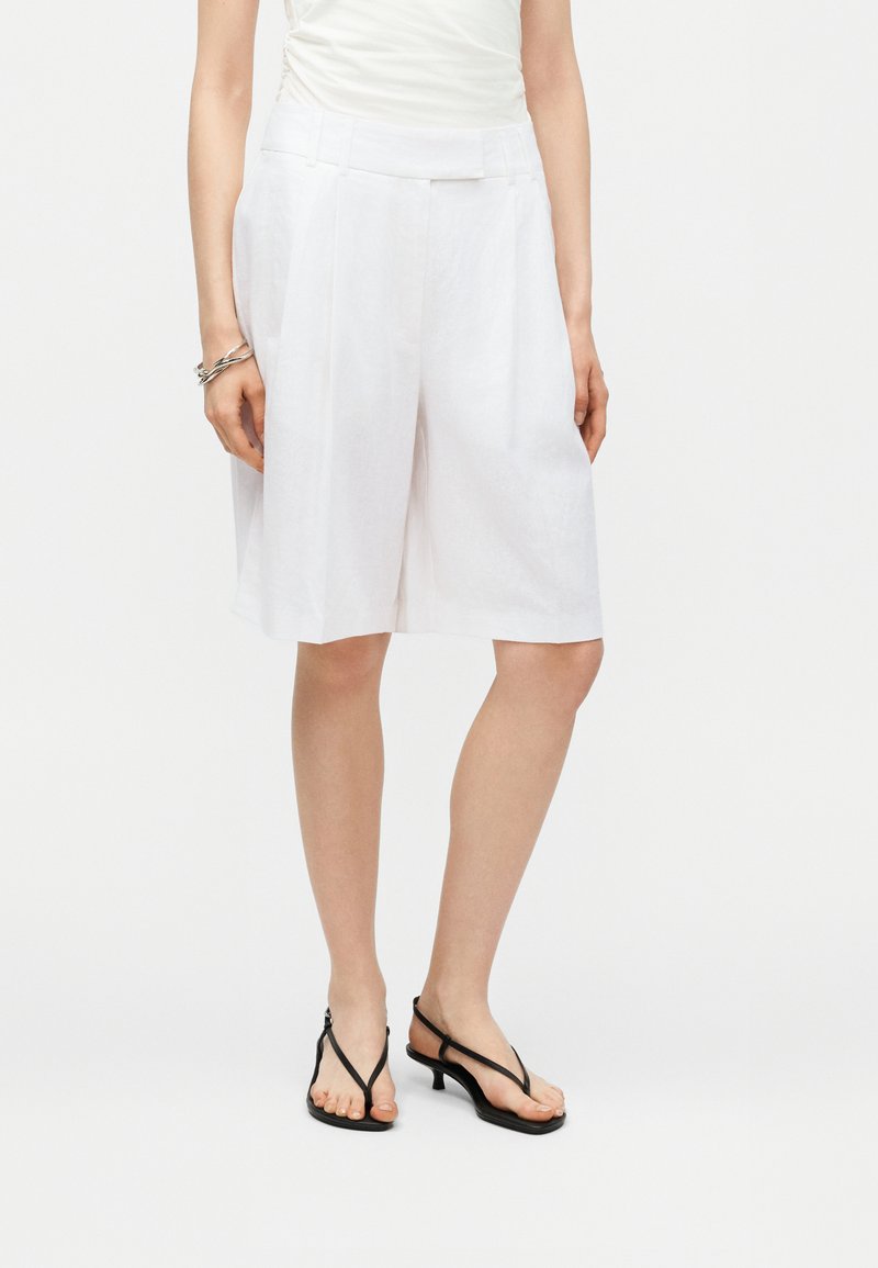 Γυναίκα που φοράει λευκή ψηλόμεση σορτς με φαρδιά πόδια μέχρι το γόνατο, λευκό αμάνικο τοπ, μαύρα σανδάλια με λεπτά λουράκια και ασημένιο βραχιόλι στον αριστερό καρπό.