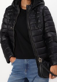 Zwarte donzen jas met een capuchon, ritssluiting en elastische trekkoorden; gestructureerde stof met een gewatteerd patroon, gecombineerd met een zwarte top en blauwe spijkerbroek.