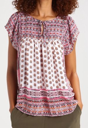 Frau trägt eine kurzärmelige Bluse mit pinken, weißen und orangefarbenen Blumenmustern und einem Schlüssellochausschnitt mit Bindeband.