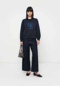Felpa navy con design grafico blu, abbinata a jeans scuri a gamba larga. La modella tiene una piccola borsa marrone e indossa scarpe scure.