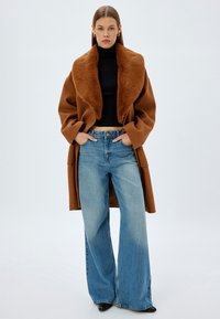 Brun ylleblandad kappa med en stor fuskpälskrage, matchad med en svart ribbad tröja och vida blå denimjeans. Svarta spetsiga skor.
