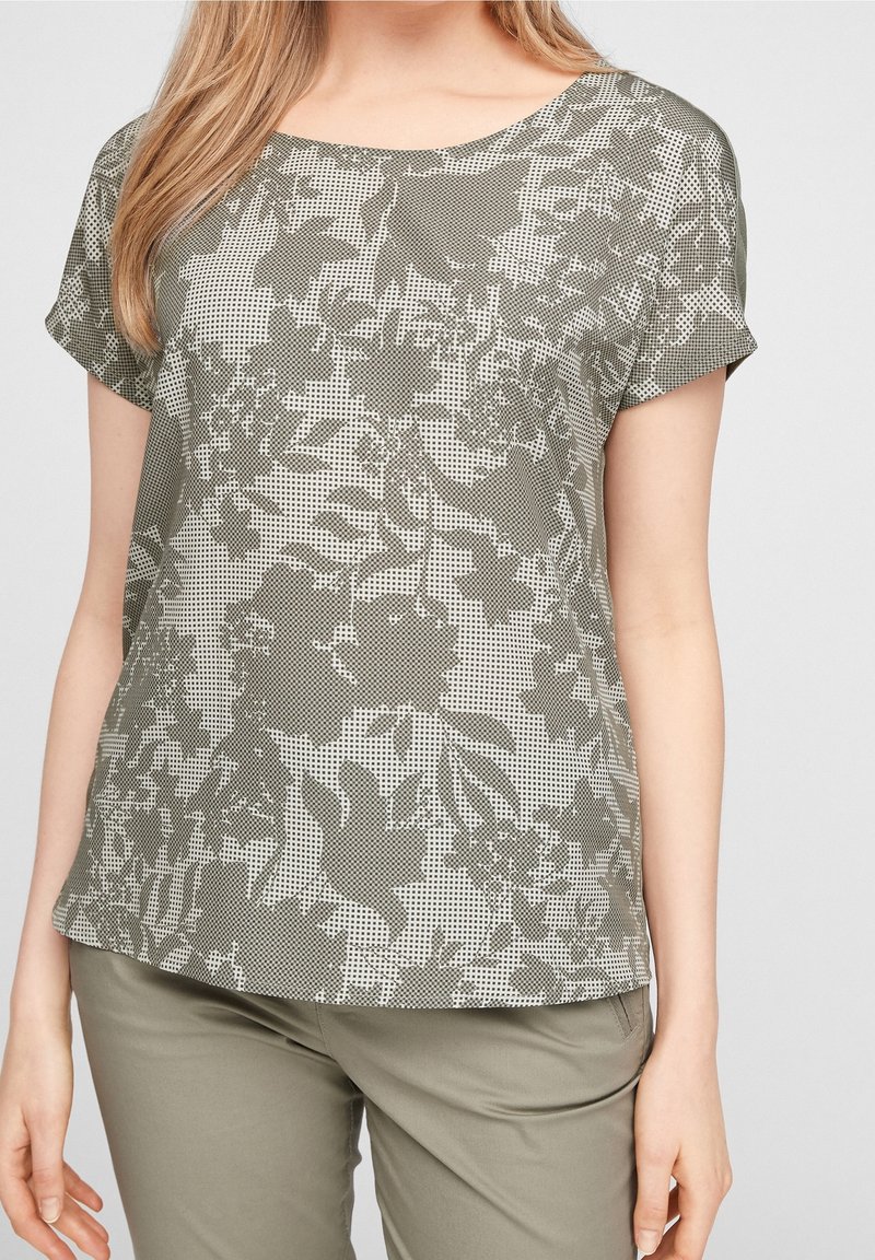 Femme portant un haut gris à manches courtes avec imprimé floral foncé et un pantalon gris clair, debout devant un fond uni.