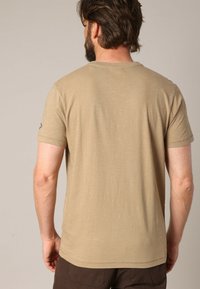 T-shirt beige à manches courtes en tissu doux, avec un col rond et une texture subtile, présentant un petit logo sur la manche gauche.