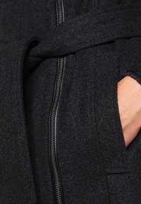 Abrigo negro de mezcla de lana con cintura ceñida, que presenta un cierre de cremallera y bolsillos laterales. La tela tiene un acabado texturizado.