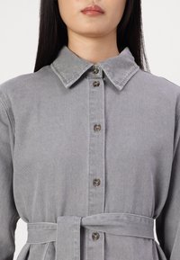 Grå denimklänning med knäppning fram, krage och midjeväska. Tyget har en slät textur med subtila sömnadsdetaljer.