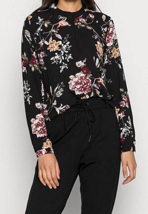 Frau trägt eine schwarze, langärmelige Bluse mit Blumenmuster in Pink, Weiß und Orange, die in eine schwarze Hose mit Tunnelzug gesteckt ist.