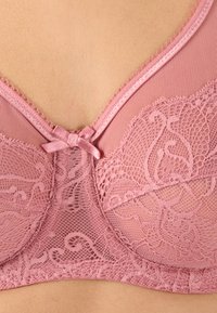 Reggiseno in pizzo di un delicato rosa con intricati motivi floreali, un fiocco centrale di raso e dettagli in rete attorno alle coppe e ai bordi.