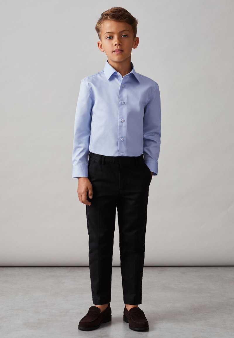 Boy standing, wearing a light blue button-up shirt, black pants, and dark loafers, against a plain gray background.