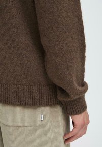 Brūns adīts džemperis ar strukturētu audumu, ar ribotām aprocēm un apakšmalu, saskaņots ar gaiši beidzādas kordūra biksēm, kuru kabatas ir ar nelielu etiķeti.