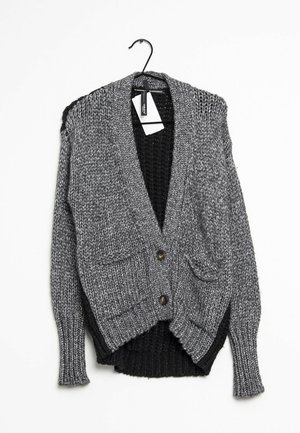 Grå chunky strikket cardigan med to knapper og forlommer, lagdelt over en sort strikket sweater på en bøjle.